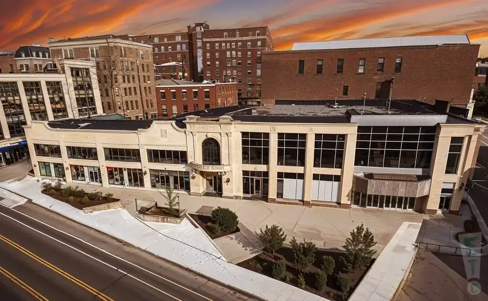 Aerial view of a light-colored building with large windows and an arched entrance, set against a dramatic orange sky.