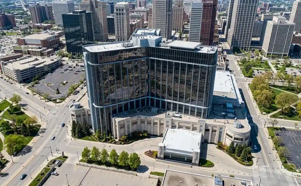 Aerial view of a large, glass-fronted building with a helipad on top, surrounded by city streets and green spaces.