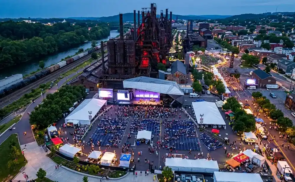 Aerial view of an outdoor concert and festival at dusk, with a stage, seating, tents, and food vendors near an industrial steel plant.