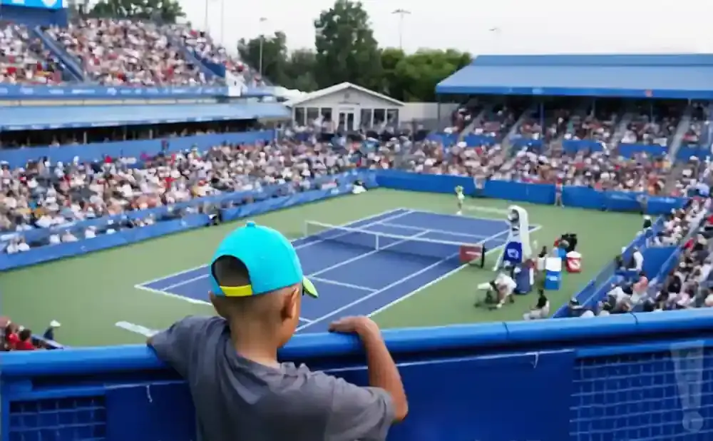A young boy in a blue cap watches a tennis match from the stands.