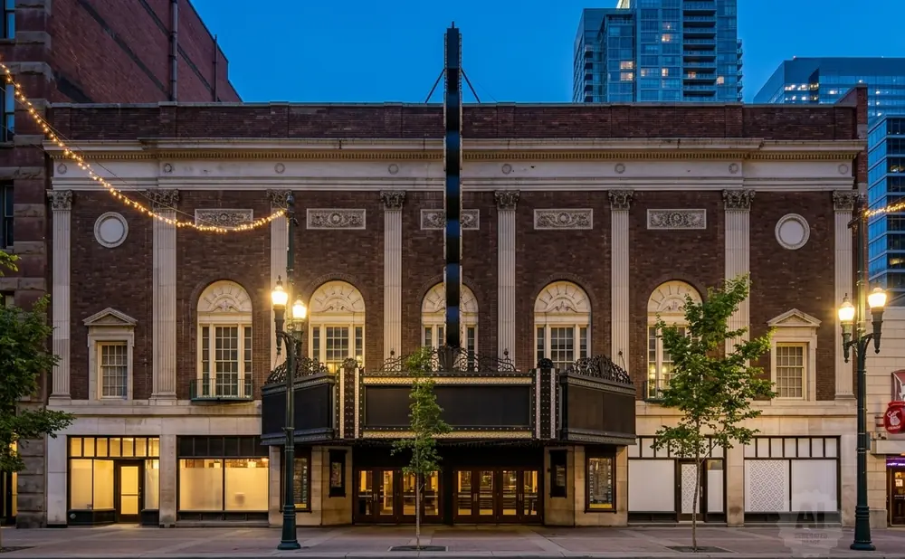 An old theater facade with a marquee, lit by strings of lights and street lamps, at dusk.