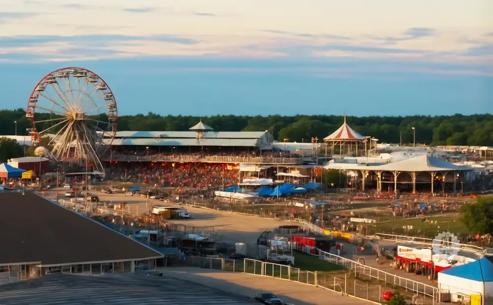 An aerial view of a busy fairground with a Ferris wheel, grandstands, and tents under a twilight sky.