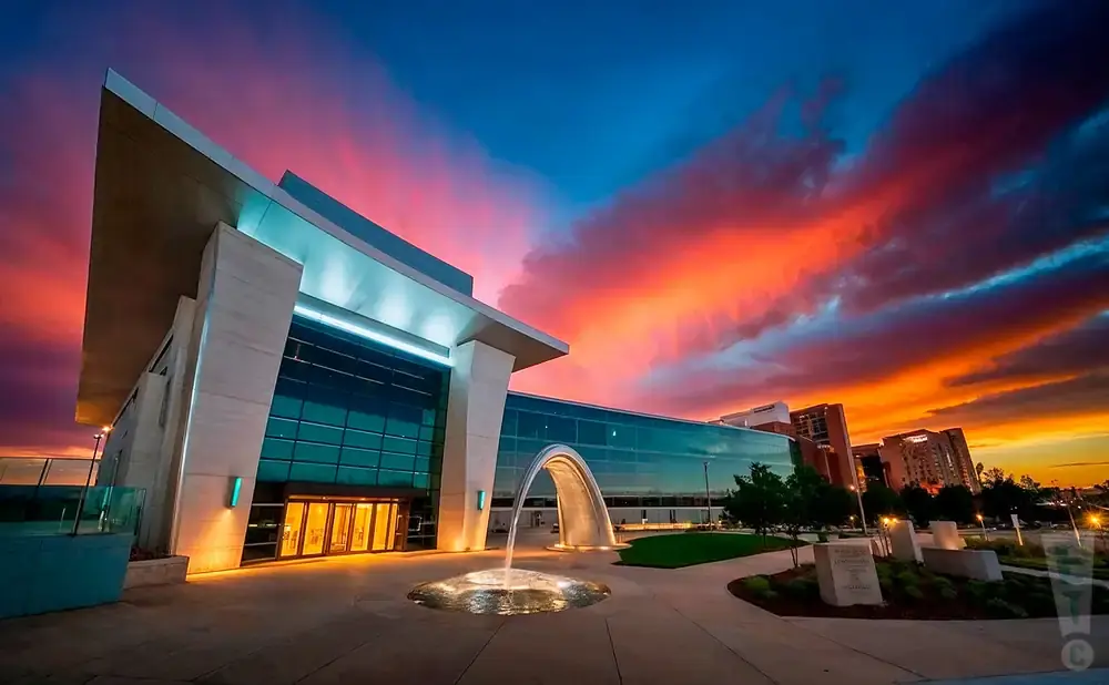 Modern building with glass facade and arched fountain at sunset with vibrant orange and blue sky.