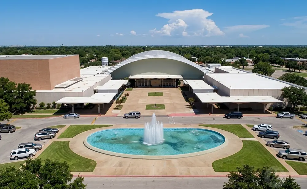 Aerial view of a large modern building with a domed roof, surrounded by trees, a fountain, and a parking lot with cars.