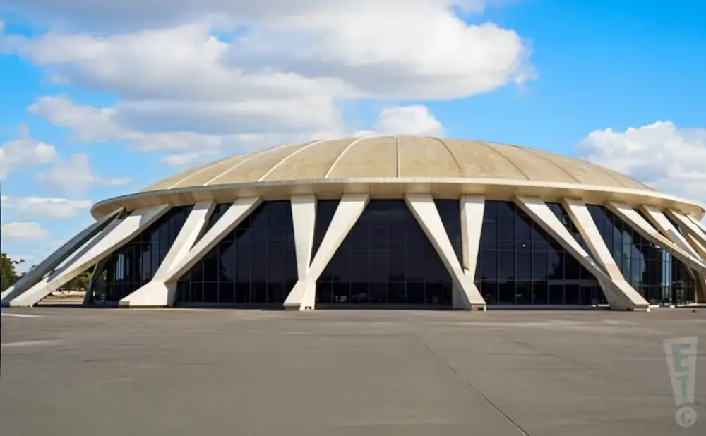 A domed building with triangular concrete supports and dark windows under a blue sky with clouds.