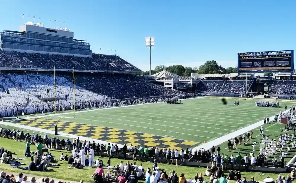 American football stadium filled with fans on a sunny day, with players on the field and goalposts visible.