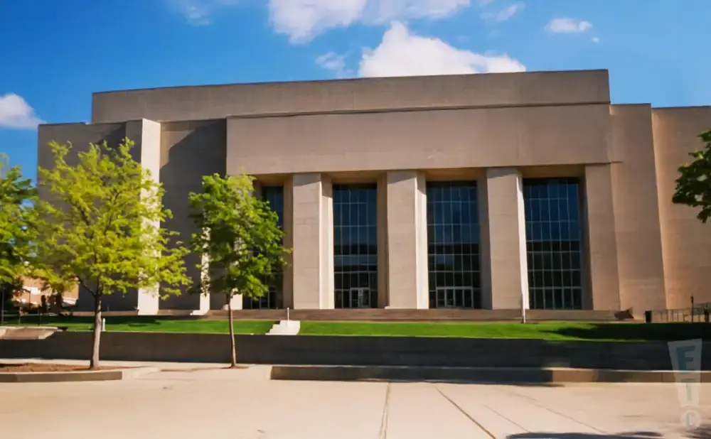 A large, tan building with tall windows and trees in front on a sunny day.