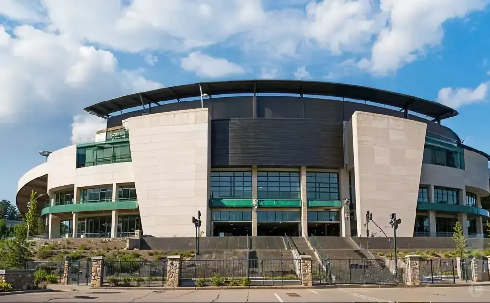 A modern stadium with a curved roof and light-colored stone facade under a blue, cloudy sky.