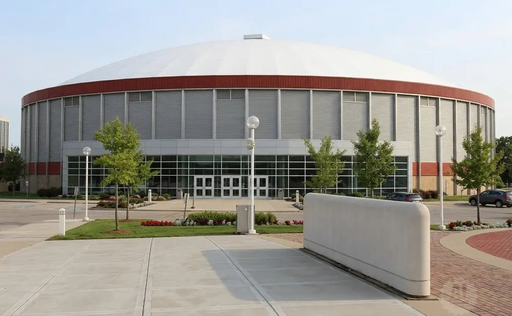 Modern domed arena with red trim and large windows, surrounded by trees and a parking lot.