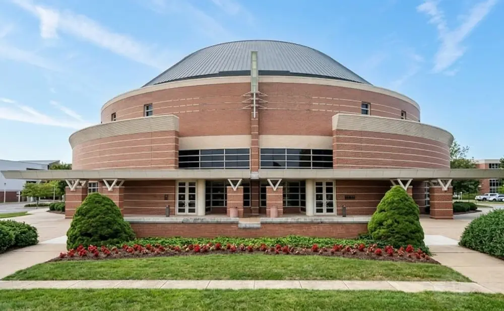 A circular brick building with a domed roof is surrounded by greenery and a blue sky.