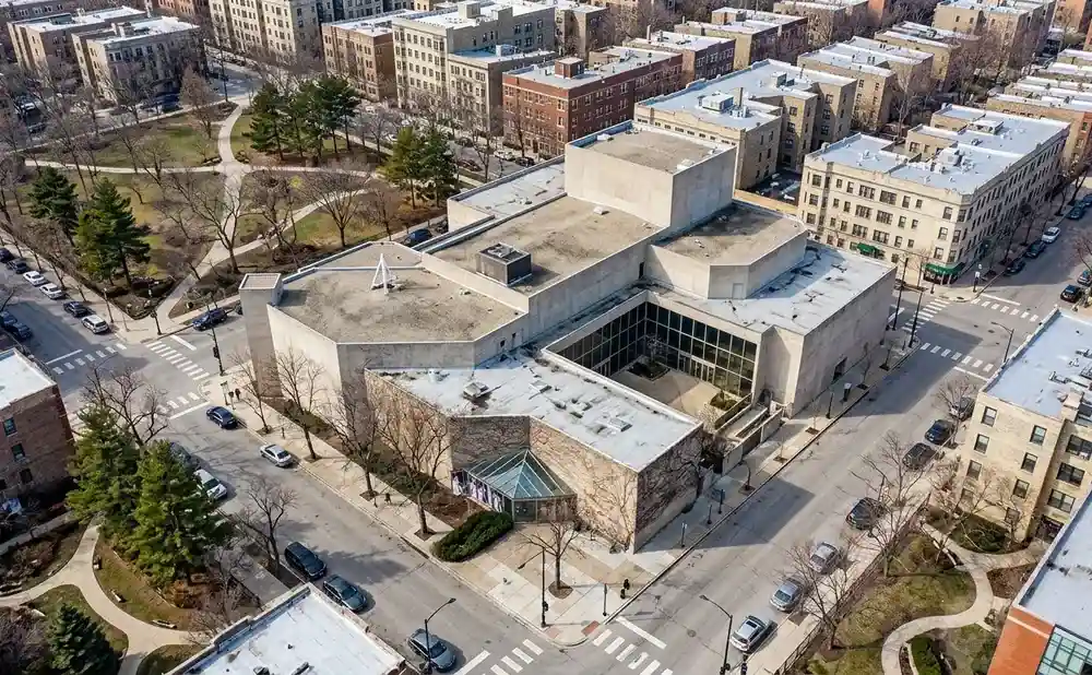 Aerial view of a city block with a large modern building at the center, surrounded by apartment buildings and a park.