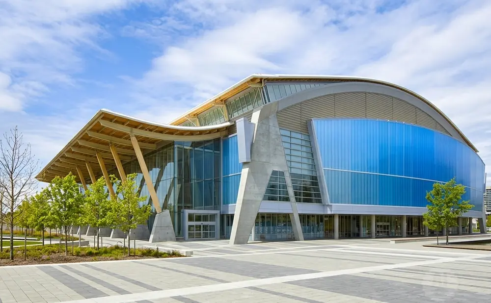 Modern building with a curved roof, glass facade, and concrete accents, surrounded by trees and a paved plaza.