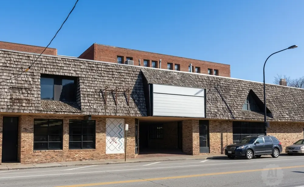 A modern brick building with a wood-shingled roof and large windows, parked cars on the street, and a clear blue sky.