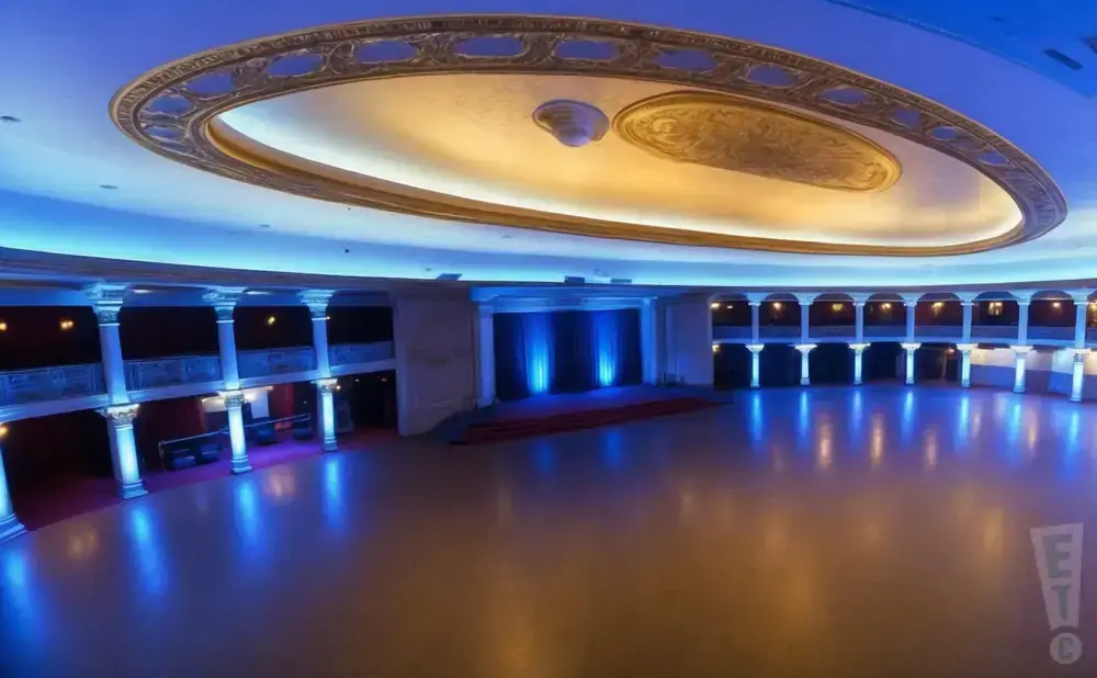 A grand ballroom with a stage, columns, and ornate ceiling, illuminated by blue lights reflecting off the floor.