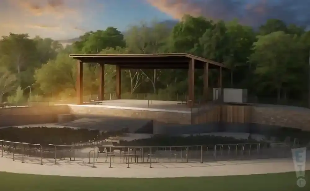 Outdoor amphitheater with a covered stage, tiered seating, and surrounding trees under a cloudy sky.