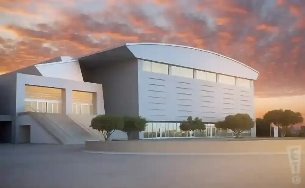 A modern, gray building with large glass entrances and windows, set against a dramatic orange and pink sunset sky.