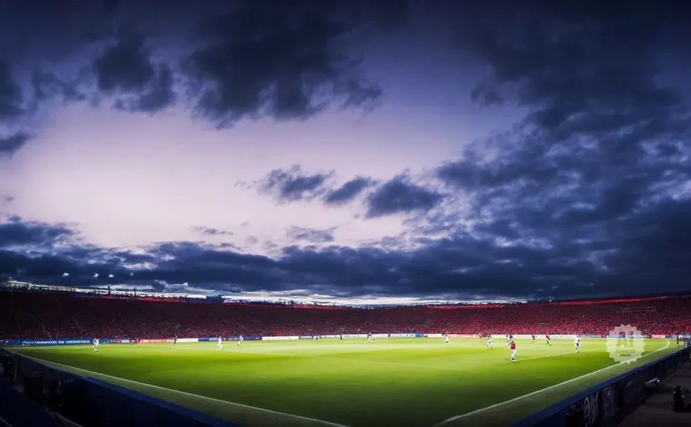 Soccer stadium at dusk with a vibrant green field, a crowd in red, and dramatic clouds in the sky.