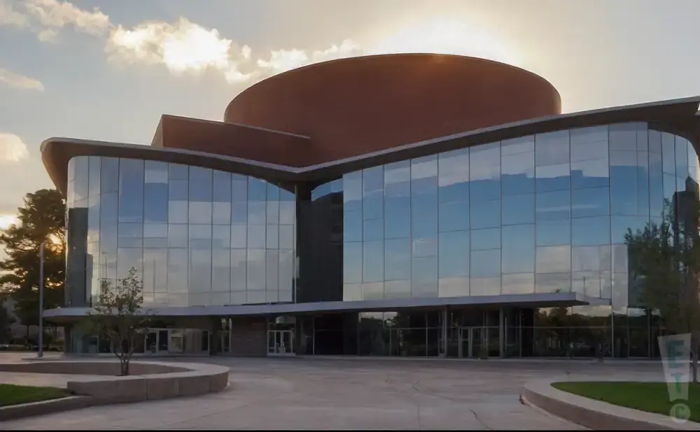 A modern glass building with a rounded, copper-colored roof against a cloudy sky at sunset.