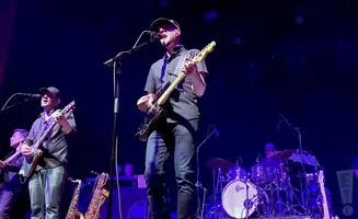 A band performs on a dimly lit stage, with a guitarist in the foreground singing into a microphone.