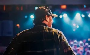A man in a cap and plaid shirt is on a stage, facing away from the camera, with a blurred audience and blue stage lights behind him.