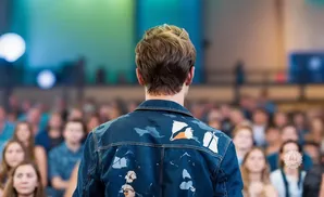 A man with wavy hair wears a denim jacket with white and orange paint splatters, facing an audience.