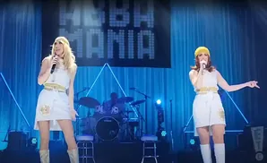 Two women in white dresses and gold boots perform on stage with 
