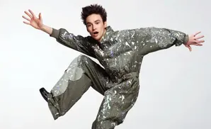 A young person in a paint-splattered jumpsuit jumps with arms outstretched against a white background.
