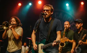 A band performs on stage with red and blue lights; a guitarist sings into a mic, a trombonist is on the left, and saxophonists are in the background.