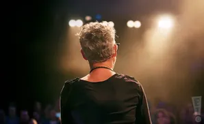 Back of a person with gray, curly hair on stage, facing a spotlight and an audience.