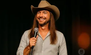 A man in a cowboy hat and a gray shirt holds a microphone and smiles.
