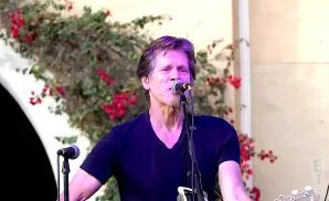 Kevin Bacon singing into a microphone while playing guitar.