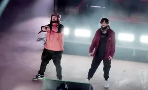 Two men perform on stage, one in an orange jacket and fur hat, the other in a red plaid shirt.