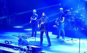 A band performs on a stage lit with blue spotlights, with singers playing guitars and a drummer in the background.