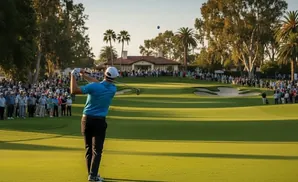 A golfer swings at a ball on a sunny course lined with spectators, palm trees, and a clubhouse.