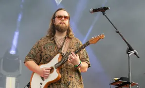 A male guitarist with long brown hair and sunglasses plays a white electric guitar on stage, with a microphone stand visible.