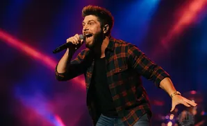 A man in a plaid shirt sings into a microphone on a stage with purple and red lights.