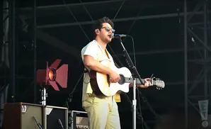 A male musician with sunglasses, a light yellow shirt, and tan pants plays an acoustic guitar and sings into a microphone on stage.