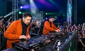 Two DJs in orange jackets perform at a concert with a cheering crowd.