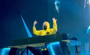 A person in a yellow hoodie and mask is standing behind a DJ booth, raising their hands.