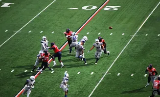 American football players in red and gray uniforms on a green field, with a football in mid-air.