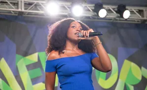 A woman with curly hair sings into a microphone on stage, wearing a blue off-the-shoulder top.