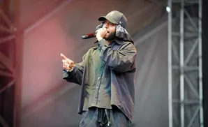 Rapper wearing a black hat and bandana, holding a microphone and gesturing on stage.