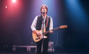 A man plays an electric guitar and sings into a microphone on a stage lit by red and blue lights.