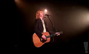 Musician with blonde hair plays an acoustic guitar and sings into a microphone while wearing a black jacket and a patterned shirt.