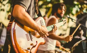 Musicians play guitars and drums outdoors, with a guitarist in the foreground playing a hollow-body electric guitar.