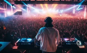 DJ with headphones on, facing a massive crowd with bright stage lights.