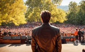 Man in leather jacket on stage facing a large outdoor audience.