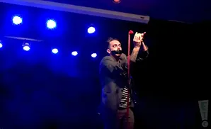 A man with black tape over his mouth stands on stage with his hands raised, holding a red ribbon.