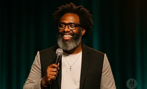 A Black man with an afro, glasses, and a beard smiles while holding a microphone.