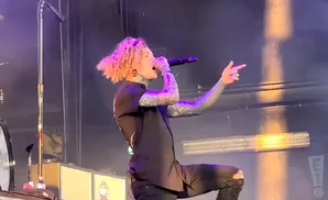 A tattooed singer with curly blonde hair sings into a microphone on stage, wearing a black shirt and ripped jeans.